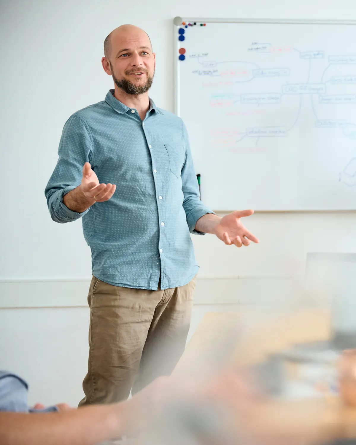 Marco Hölzel erklärt am Whiteboard ein Entwicklungskonzept, im Hintergrund arbeitet ein Mitarbeiter am Stereomikroskop