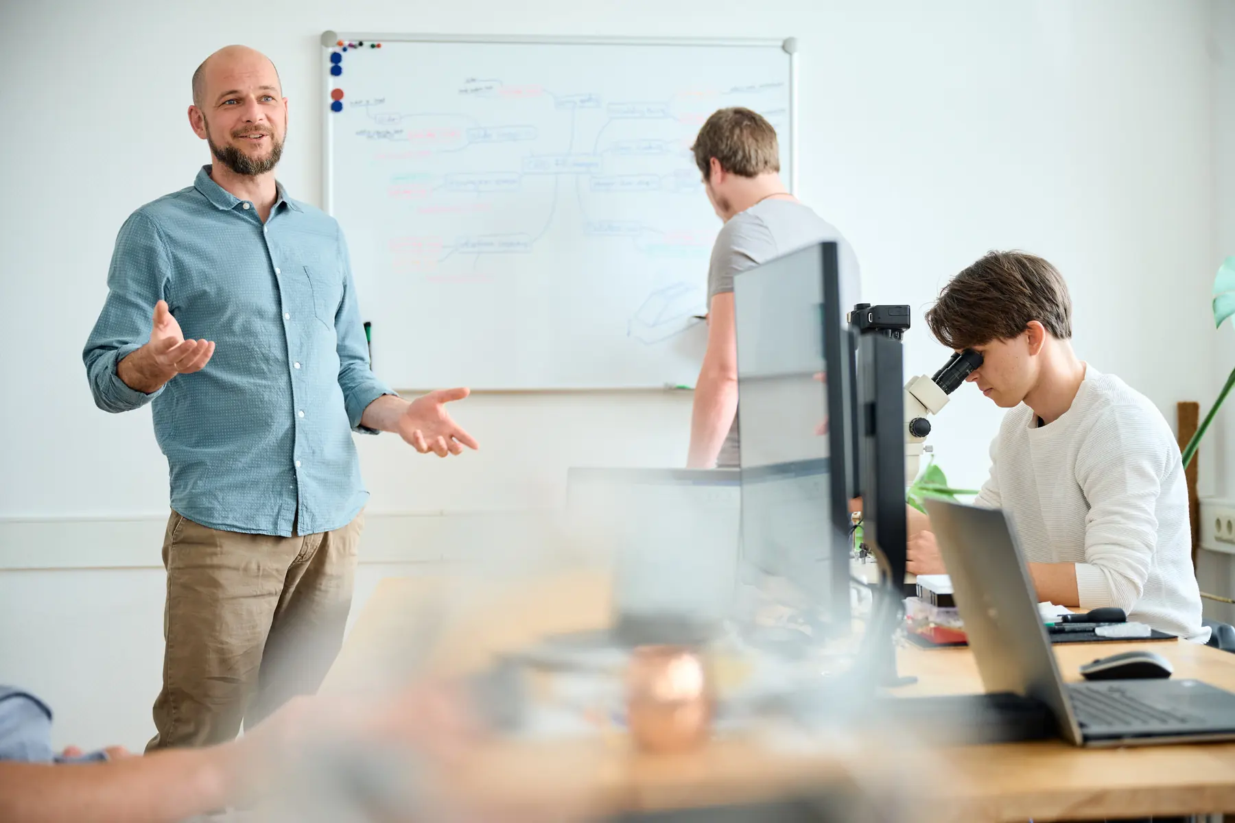 Marco Hölzel erklärt am Whiteboard, ein Kollege arbeitet daneben, ein Mitarbeiter schaut durch ein Stereomikroskop